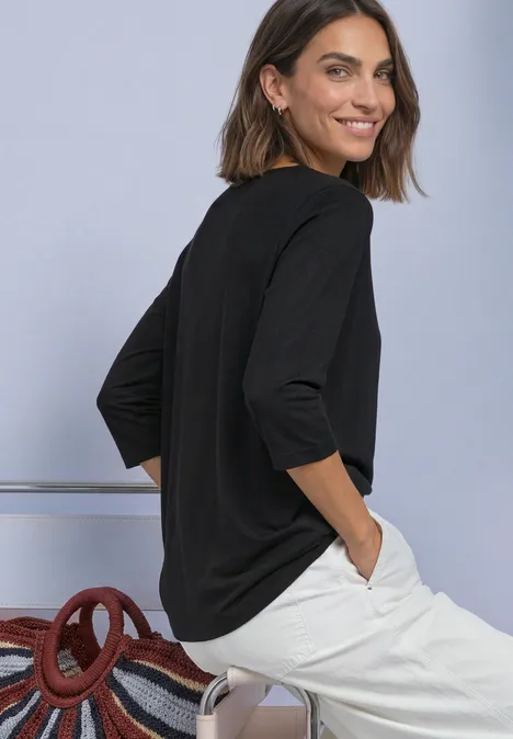 Shirt met 3/4-mouwen en een boothals Black Frau im schwarzen 3/4-Arm-Shirt und weißen Hosen sitzt seitlich gedreht, lächelnd und mit Händen in den Taschen.