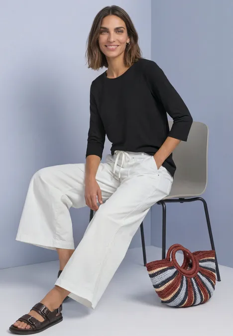 Shirt met 3/4-mouwen en een boothals Black Frau in schwarzem Oberteil, weißen Culotte-Hosen und Sandalen sitzt auf Stuhl, bunte Tasche daneben.