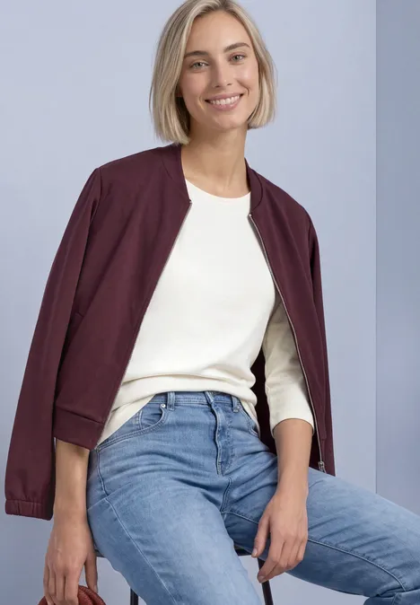 Frau mit hellem Haarschnitt trägt weiße Bluse und Jeans, eine weinrote Jacke locker über den Schultern, sitzt lächelnd.