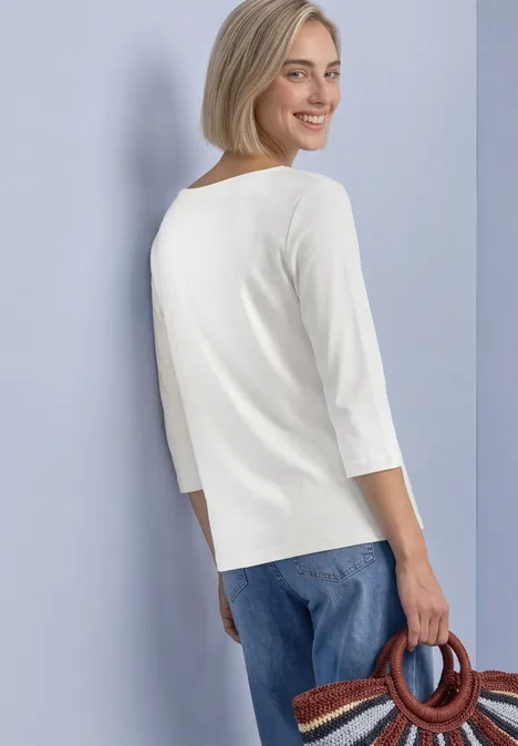 Frau im Profil, Blick über die Schulter, trägt ein weißes 3/4-Arm-Shirt, blaue Jeans und eine runde Flechttasche.