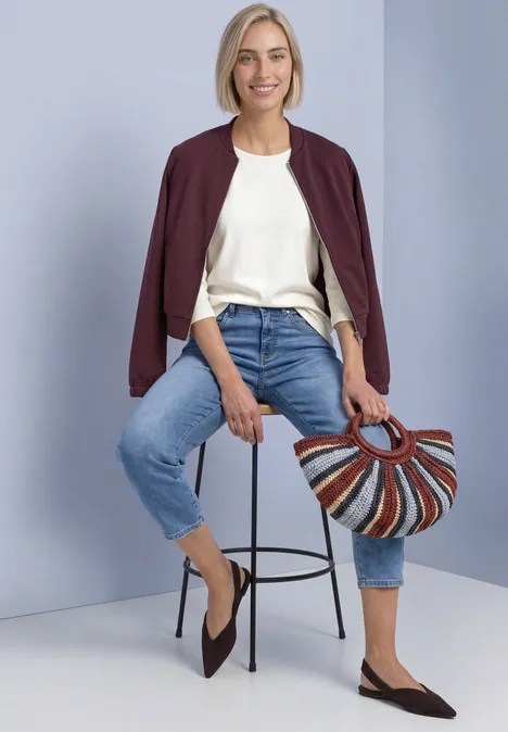 Frau in weißem Shirt, Jeans und burgunderrotem Jäckchen sitzt auf einem Hocker und hält eine bunte Korbtasche.