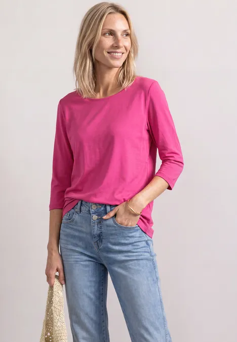 Een lachende vrouw in een roze 3/4-mouwshirt en jeans, met een gevlochten tas in haar hand.