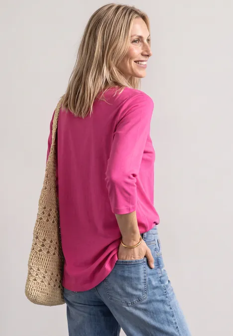 Vrouw in roze shirt met driekwartmouwen, spijkerbroek en gehaakte tas, zijwaarts staand en omkijkend.