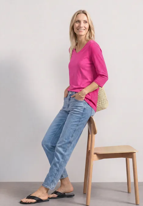 Vrouw in roze top, spijkerbroek en zwarte sandalen leunt ontspannen tegen een kruk voor een witte achtergrond.