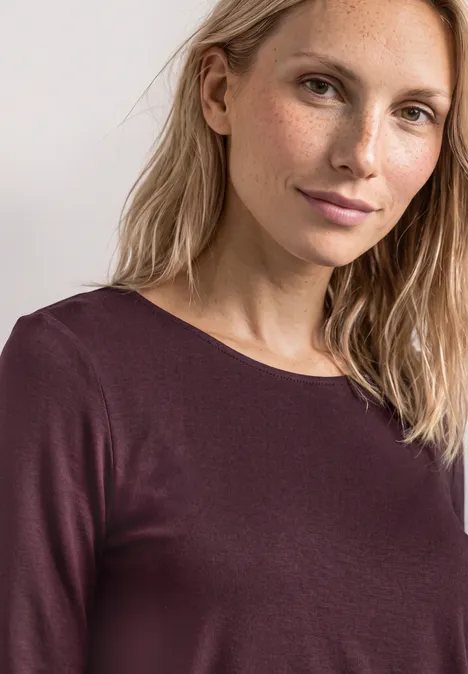 Frau mit blonden Haaren und dezentem Lächeln in einem dunkelvioletten Langarmshirt vor hellem Hintergrund.