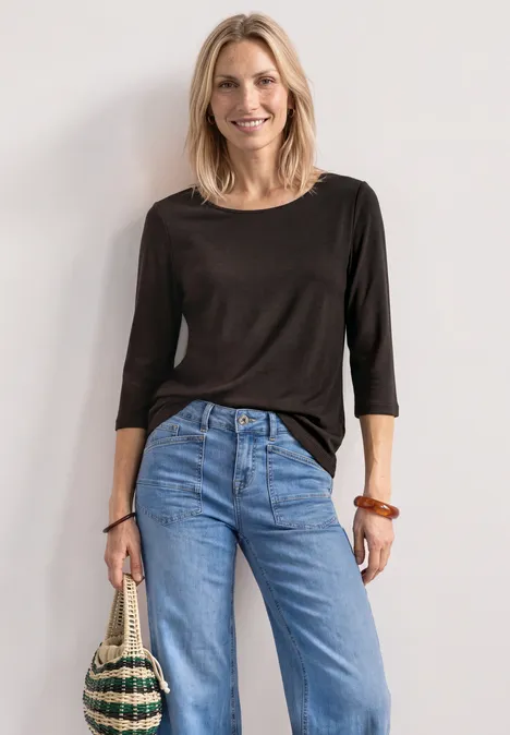 Shirt met 3/4-mouwen en een boothals black coffee Lachende vrouw in zwart topje en blauwe jeans, met een strooien tas voor een lichte achtergrond
