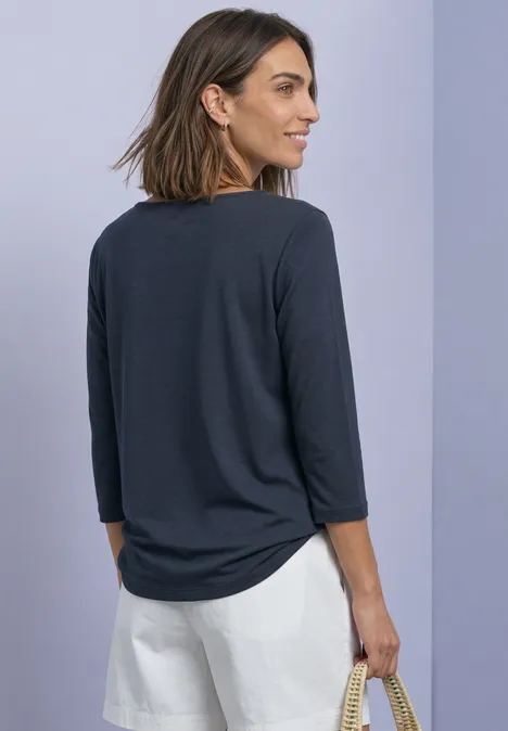 Shirt met 3/4-mouwen en een boothals darkest blue Frau in dunkelgrauem 3/4-Arm-Shirt und weißen Shorts, seitlich gewandt mit Korbtasche in Hand vor lila Hintergrund.