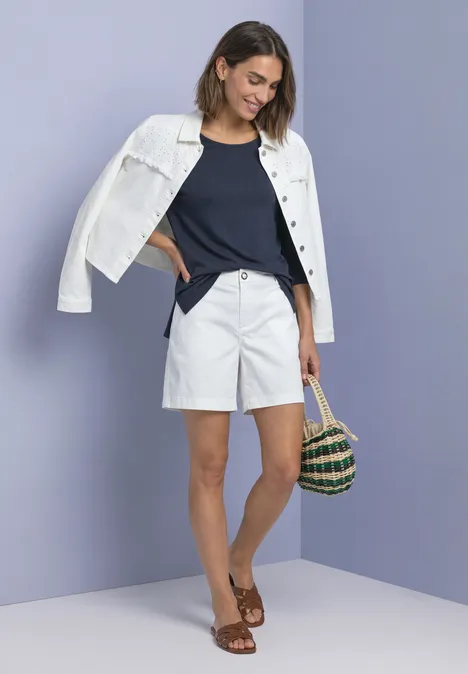 Shirt met 3/4-mouwen en een boothals darkest blue Frau mit weißer Jacke auf den Schultern, dunkelblauem Top, weißen Shorts, braunen Sandalen und Korbtasche vor lila Wand.