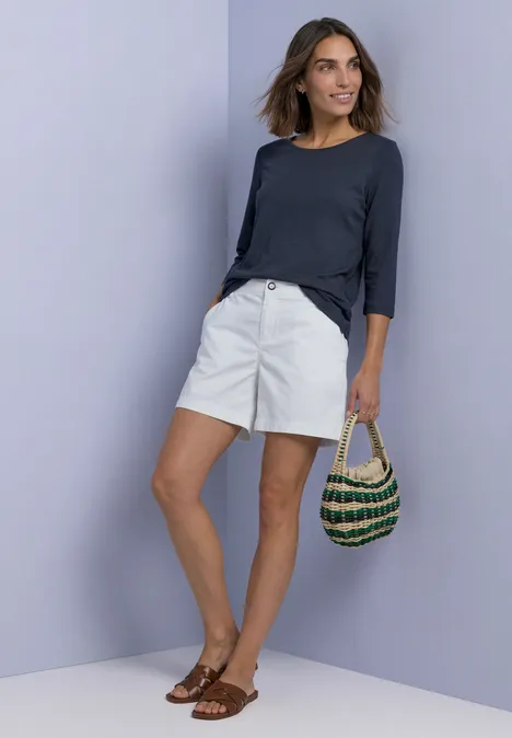 Shirt met 3/4-mouwen en een boothals darkest blue Frau in weißer Shorts, dunkelblauem Oberteil, Sandalen, hält einen gehäkelten Korb vor pastellfarbener Wand.