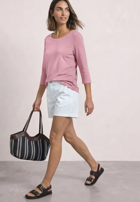Frau in rosa Shirt und weißen Shorts mit gestreifter Tasche und schwarzen Sandalen vor hellem Hintergrund.