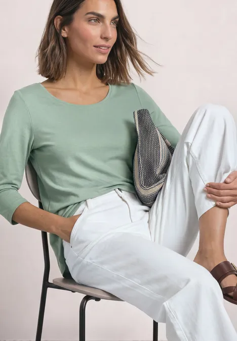 Frau mit mittellangem Haar sitzt auf Stuhl, trägt mintgrünes 3/4-Arm-Shirt, weiße Hose und braune Sandalen.