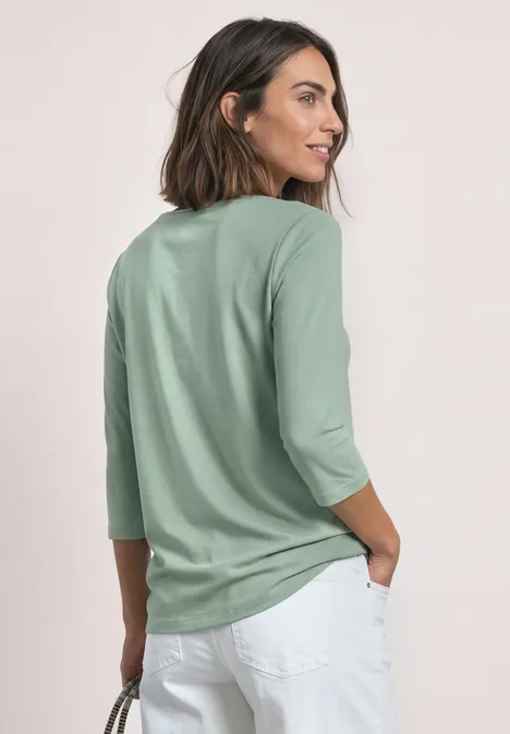 Frau von hinten in mintgrünem Shirt mit 3/4-Ärmeln und weißen Hosen vor neutralem Hintergrund.