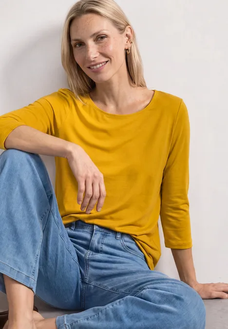 Lächelnde Frau mit blonden Haaren, gelbem Shirt und Jeans sitzt entspannt auf dem Boden vor weißer Wand.
