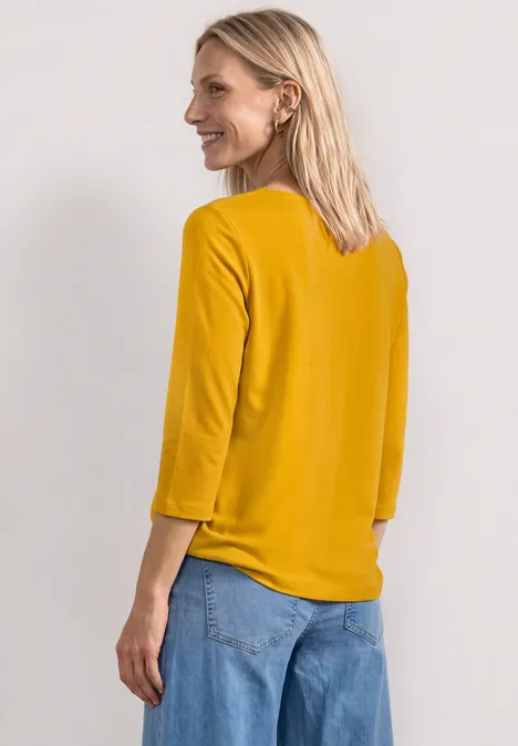 Eine Frau von hinten in senfgelbem Oberteil mit 3/4-Ärmeln und blauer Jeans.