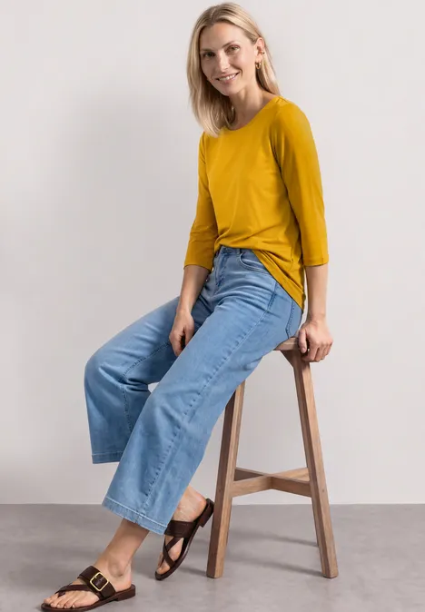 Frau sitzt auf Hocker, trägt senfgelben 3/4-Arm-Pullover, weite Jeans und schwarze Sandalen.