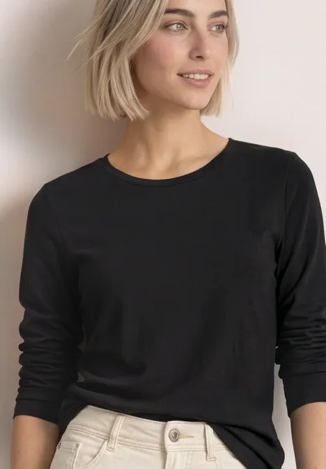 Blonde Frau im schwarzen Langarmshirt und hellen Hosen vor schlichtem Hintergrund.