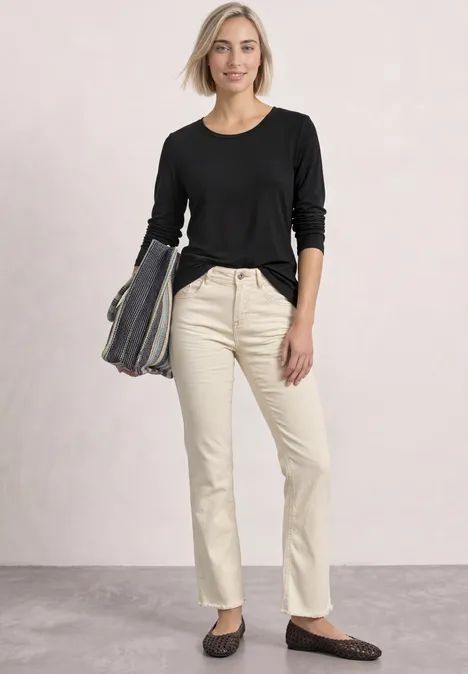 Eine lächelnde Frau in schwarzem Langarmshirt und beigen Jeans, mit Tasche in der Hand.
