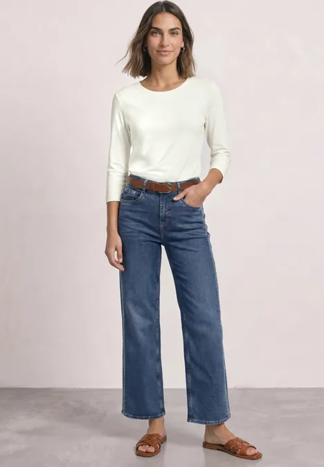 Frau steht vor hellem Hintergrund, trägt weißes Langarmshirt, blaue Jeans, braunen Gürtel und Sandalen.
