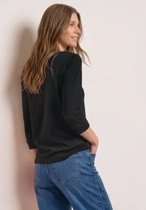 3/4-Arm Shirt mit U-Boot-Ausschnitt Black Eine Frau von hinten in schwarzem Oberteil und blauen Jeans, die entspannt an einer Wand steht.