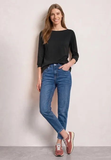 3/4-Arm Shirt mit U-Boot-Ausschnitt Black Eine Frau in schwarzem Langarmshirt, blauen Jeans und Sneakers steht lässig vor einer hellen Wand.