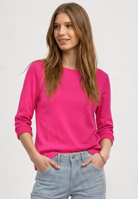 Junge Frau mit langem braunem Haar trägt pinken Pullover und Jeans, lächelt vor neutralem Hintergrund.