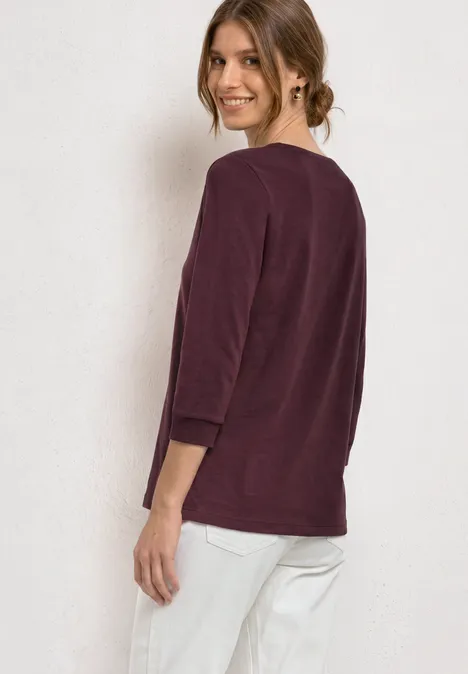 Frau in dunkelrotem 3/4-Arm-Shirt und weißen Hosen, seitlich stehend, Blick nach hinten.
