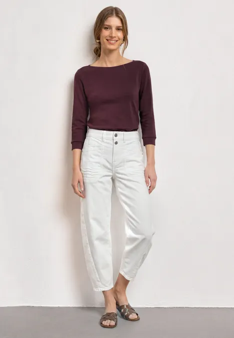 Frau in bordeauxrotem Langarmshirt und weißen, weiten High-Waist-Jeans mit grauen Sandalen vor weißem Hintergrund.