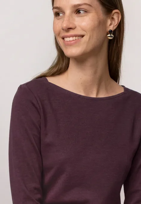 Eine junge Frau mit langem, braunem Haar trägt bordeauxfarbenes Shirt und goldene Ohrringe.