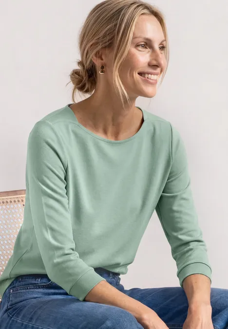 Frau in mintgrünem Pullover und Jeans sitzt entspannt seitlich auf einem Stuhl und blickt zur Seite