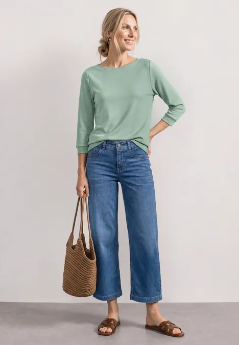Frau in grünem 3/4-Shirt, weiten Jeans, braunen Sandalen und Stroh-Tasche vor neutralem Hintergrund.