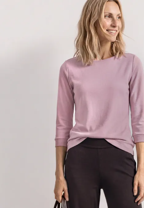 Frau in pastellrosa Dreiviertelarm-Shirt und dunkler Hose vor hellem Hintergrund