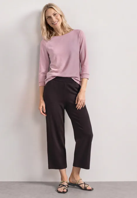 Eine Frau trägt rosa Langarmshirt, schwarze weite 7/8-Hose und schwarze Sandalen vor hellem Hintergrund.