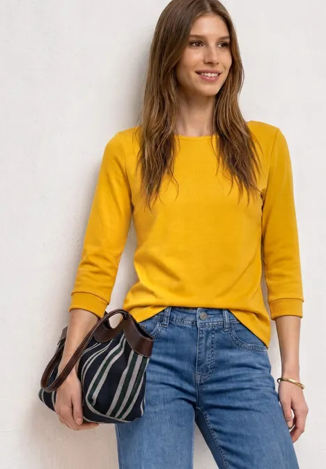 Frau trägt gelbes 3/4-Arm-Shirt, blaue Jeans und hält eine schwarz-weiße gestreifte Tasche.