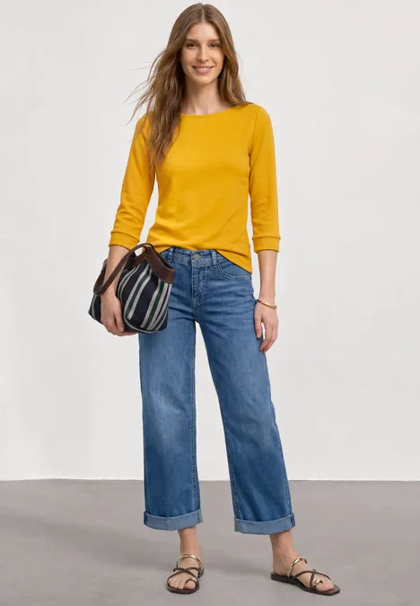 Frau in gelbem Langarmshirt, weiter Cropped-Jeans, Sandalen und gestreifter Schultertasche vor schlichter Wand.