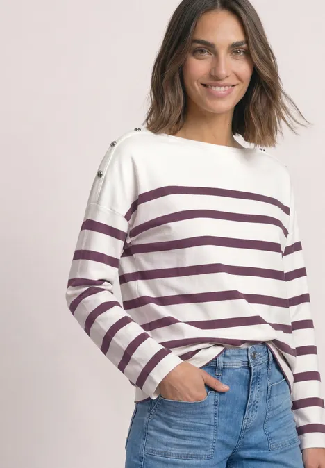 Frau in weißem Langarmshirt mit violetten Streifen und blauen Jeans, lächelnd, Hand in Tasche.