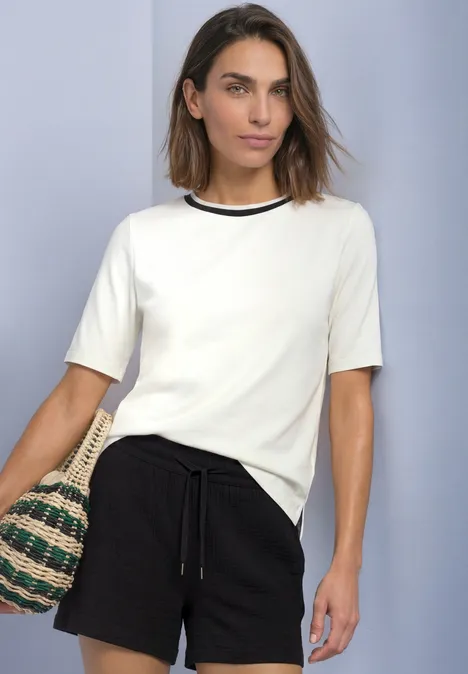T-Shirt mit Rundhals und Kontrastdetail off white Frau in weißem T-Shirt und schwarzen Shorts trägt Korbhandtasche vor einfachem Hintergrund.
