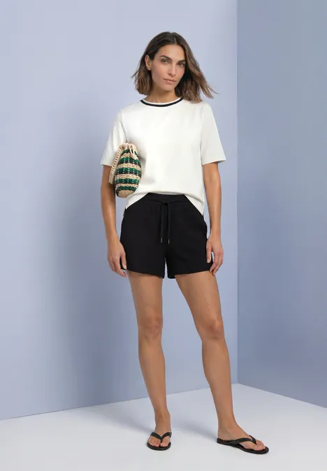 T-Shirt mit Rundhals und Kontrastdetail off white Frau trägt weißes T-Shirt, schwarze Shorts, Flip-Flops und eine geflochtene Tasche vor lila Hintergrund.
