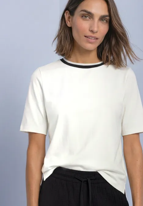 T-Shirt mit Rundhals und Kontrastdetail off white Frau in weißem Kurzarmshirt mit schwarzem Rundhals und schwarzer Hose vor neutralem Hintergrund.