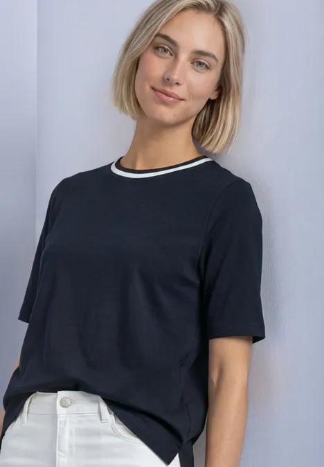 Frau mit kurzen blonden Haaren trägt schwarzes T-Shirt und weiße Jeans vor hellem Hintergrund.