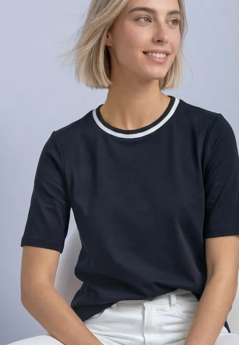 Junge Frau sitzt, blickt lächelnd seitlich, trägt schwarzes T-Shirt mit weißem Doppelkragen und weiße Jeans.