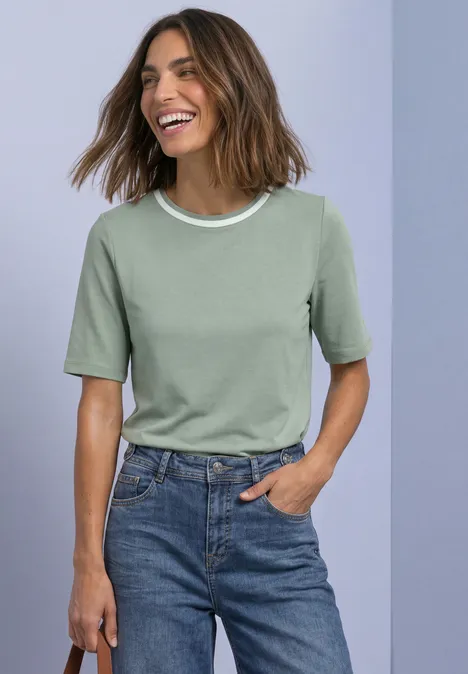 Shirt met ronde hals en contrasterende details eucalyptus green Frau lächelt, trägt grünes T-Shirt mit weißem Kragen und Jeans, steht mit einer Hand in der Tasche.