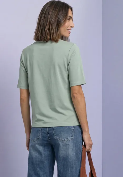 Shirt met ronde hals en contrasterende details eucalyptus green Frau von hinten in grünem T-Shirt und blauer Jeans, hält Tasche vor lila Wand.