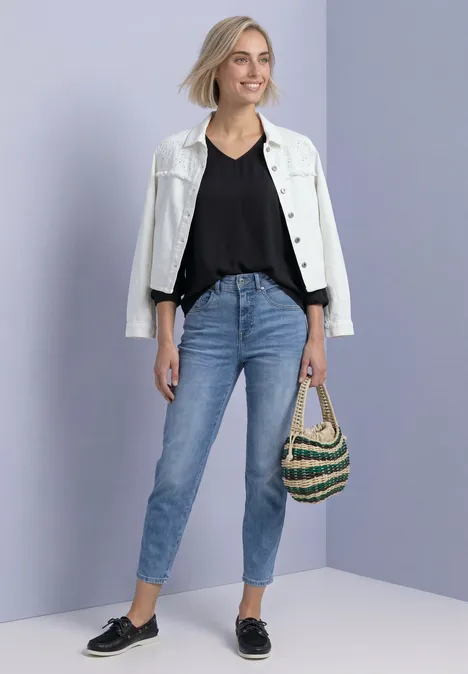 Junge Frau mit weißer Jacke, schwarzem Top, blauen Jeans, schwarzen Loafers und gestreifter Strandtasche.