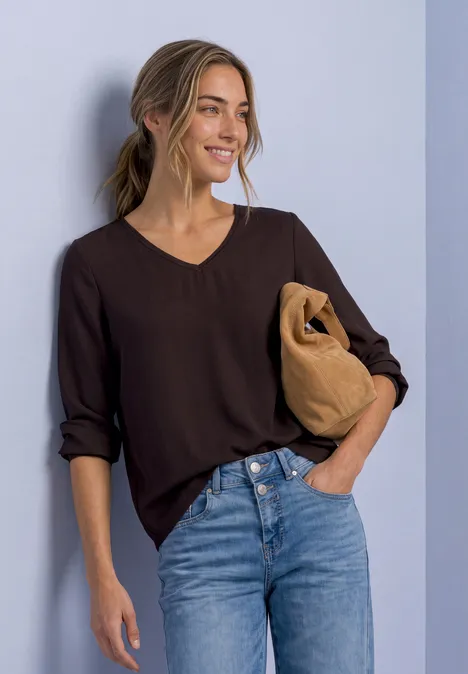 Frau in dunkelblauem V-Neck-Shirt und Jeans, lächelt, hält eine beige Tasche in der Armbeuge.