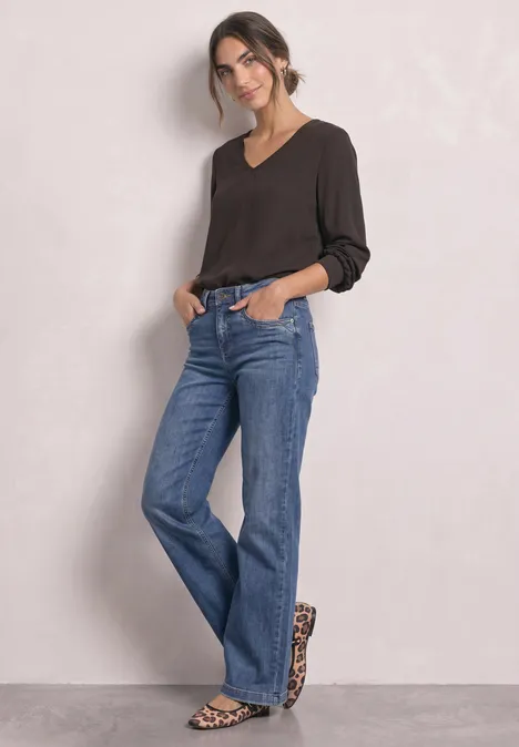 Frau in schwarzem Shirt und weiten Jeans mit Leoparden-Sandalen steht entspannt vor hellem Hintergrund.