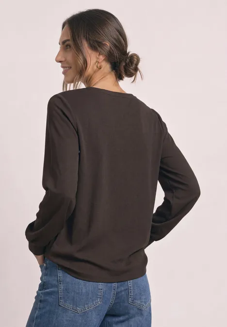 Frau im Profil trägt braunes Langarmshirt und blaue Jeans, Haare zu Dutt gebunden.