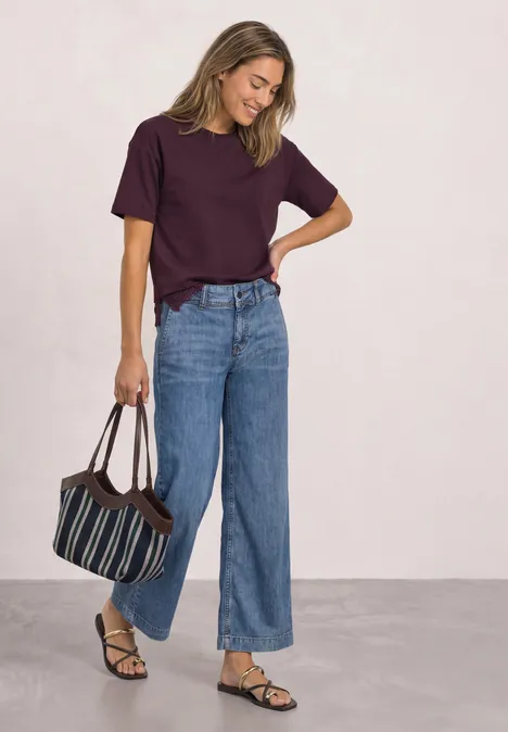 Frau in weitem Jeans, bordeauxrotem T-Shirt und Sandalen, trägt gestreifte Umhängetasche.