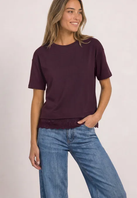 Eine junge Frau trägt ein dunkellila T-Shirt und blaue Jeans und lächelt locker.
