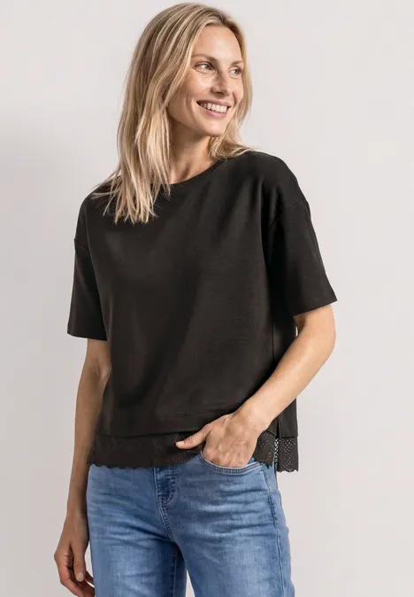Eine lächelnde Frau in schwarzem T-Shirt und blauen Jeans vor hellem Hintergrund.