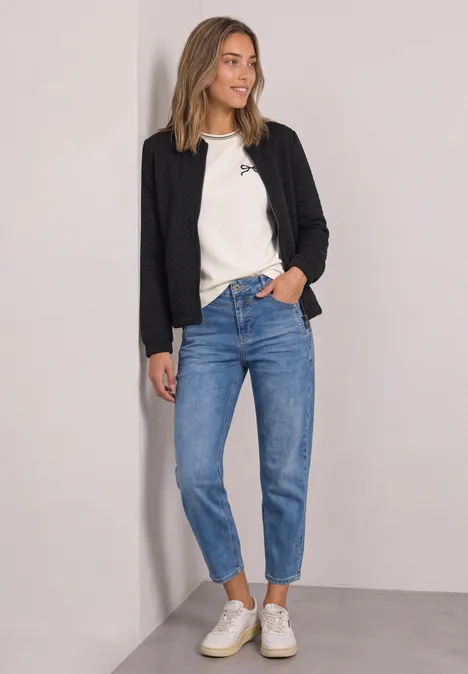 Jasje met ronde hals en structuur Black Vrouw in zwarte blazer, wit shirt, blauwe jeans en witte sneakers leunt tegen een muur.